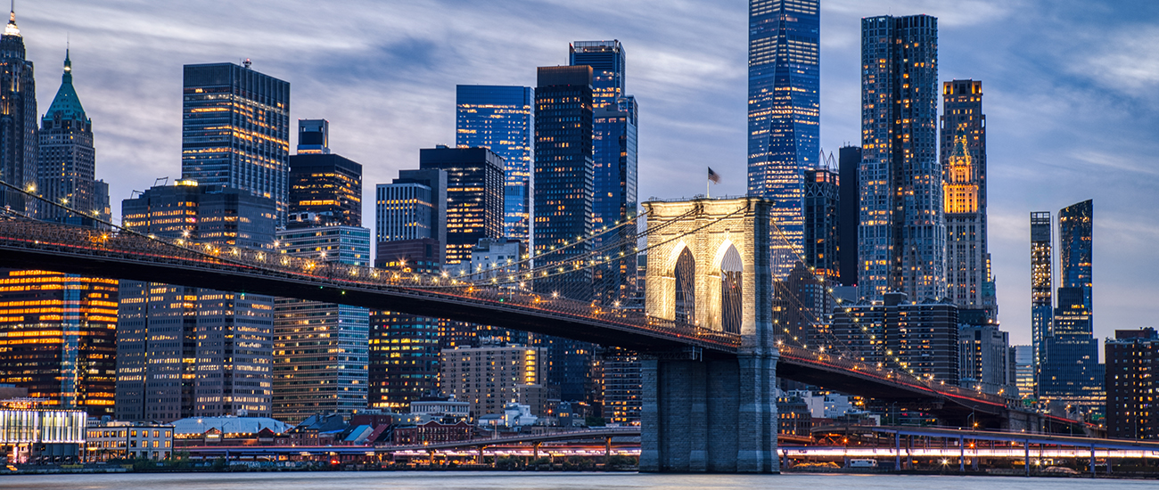 Brooklyn Bridge in New York City at dusk, illuminated with golden lights, spanning the East River against a backdrop of lit skyscrapers.